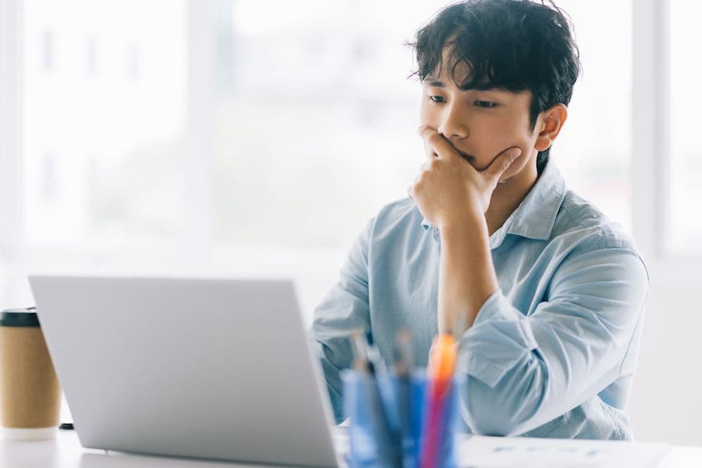 person sitting with a furrowed brow, anxiously holding his mouth as he awaits the completion of a processing task on his laptop