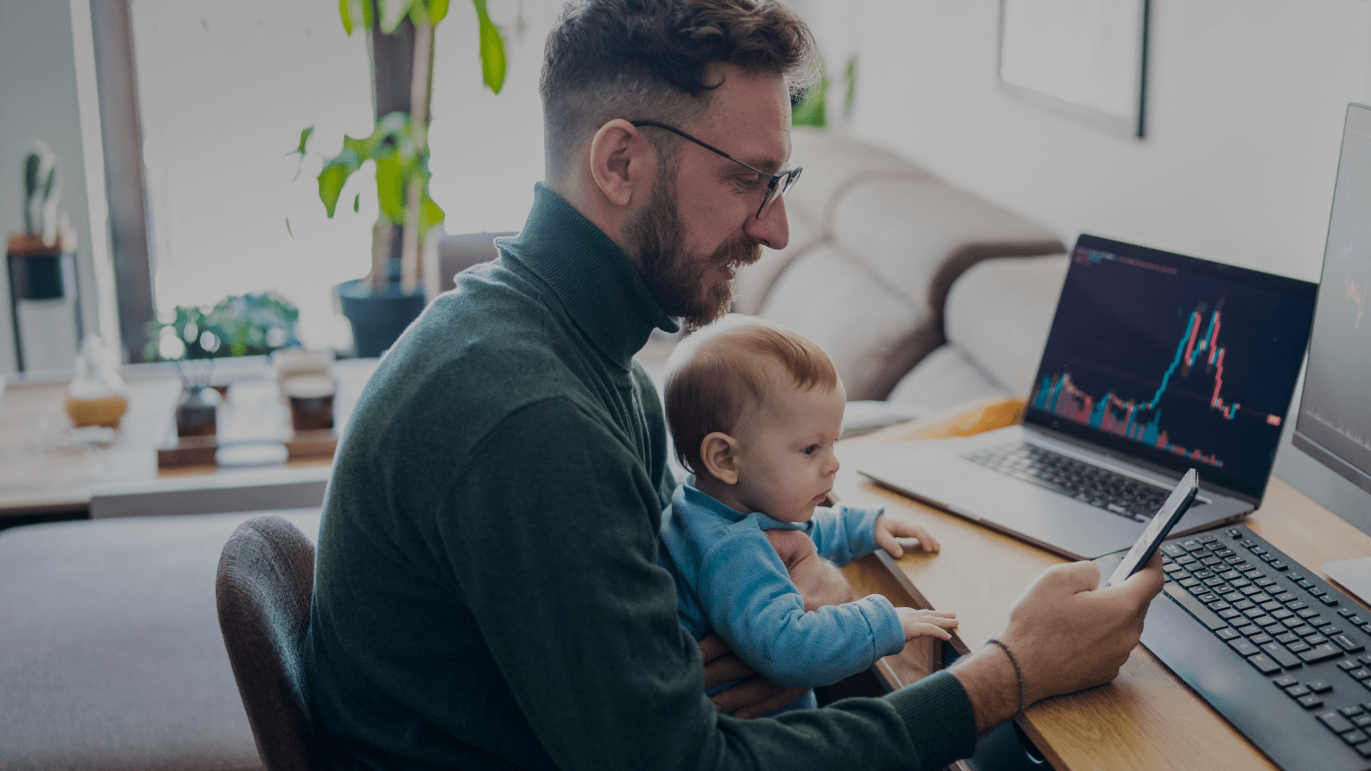 A man holds a child while looking at a phone and analyzing a financial ticker in a well-kept home office.
