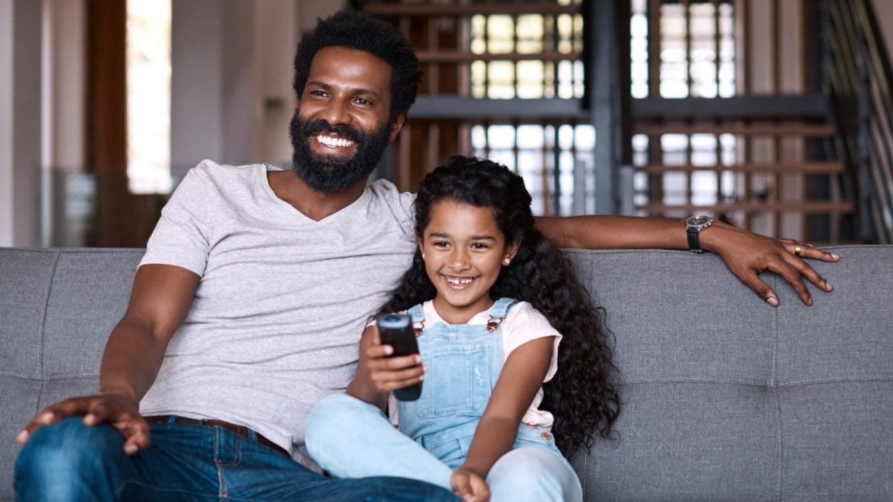 Father and his daughter sit on the sofa, happily sharing a remote control while beaming with joy
