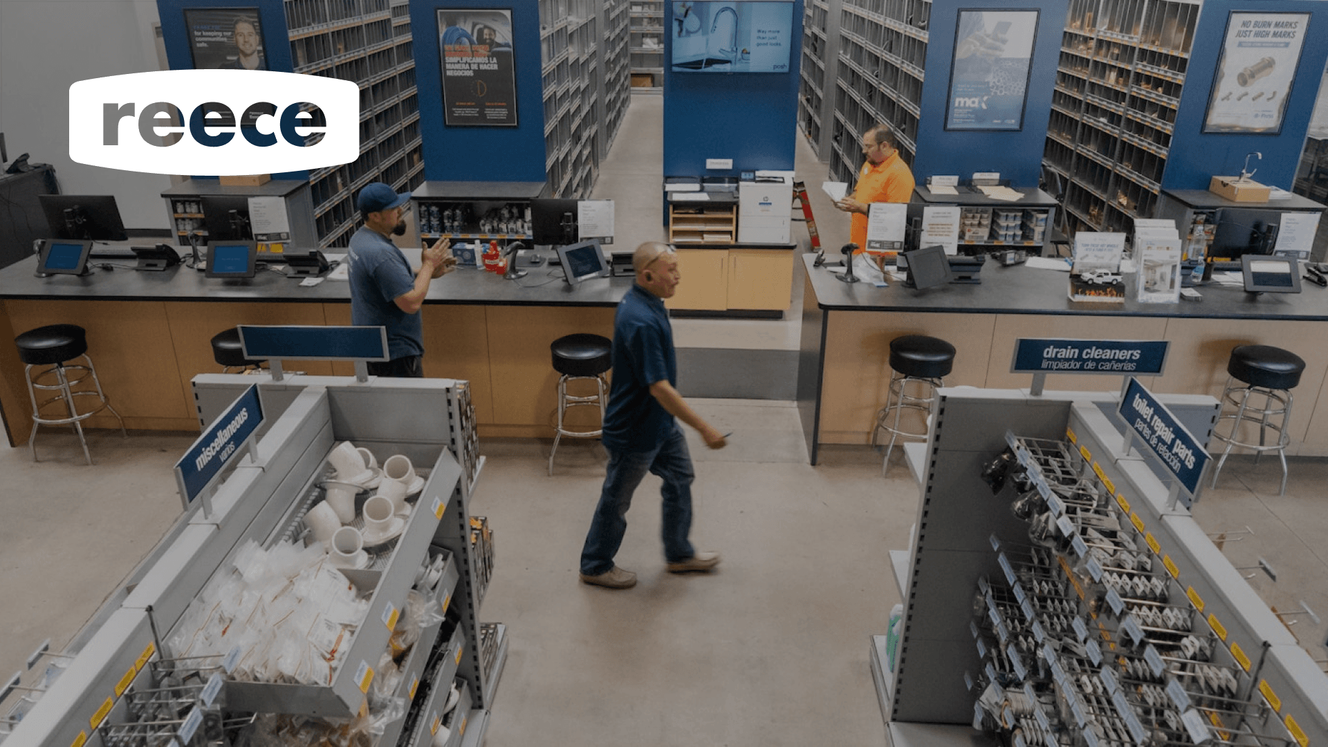 A tradesperson walks along an aisle of plumbing supplies, with the reece logo in the top left corner