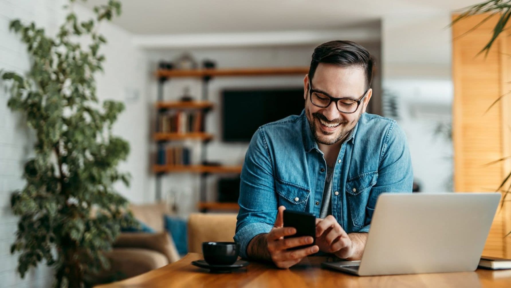 Man seated at the table with a cup and cup, multitasking as he operates his smartphone